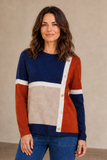 A woman wearing an Emreco color-block knit sweater with navy blue, rust orange, beige, and off-white panels, paired with blue jeans, standing against a neutral beige wall.