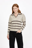 A woman modeling a MUDFLOWER beige knit sweater with dark brown horizontal stripes, a scalloped V-neckline and collar, paired with black pants, against a plain white studio background.