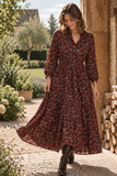 A woman wearing a BRAKEBURN long-sleeved, button-front maxi dress in an autumnal berry floral pattern, walking outdoors on a stone path next to a rustic stone wall. Available from Mooreheads, Bessbrook 