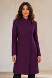 A woman modeling a long Emreco wrap-style coat in a rich deep purple color, standing against a neutral beige textured wall.