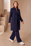 A woman models an EMRECO midnight blue single breasted revere collar coat, matching top, wide-leg trousers and white sneakers in a minimalist beige architectural setting. The coat is available to buy from W. MOOREHEAD, Bessbrook.