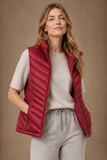 A woman wearing an Emreco sleeveless quilted puffer vest in deep burgundy red over a light beige t-shirt and matching sweatpants, posed against a solid warm brown studio backdrop.