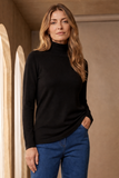 A woman wearing a black long-sleeve roll neck top and blue jeans, standing in a warm-toned indoor setting with beige walls and arched light openings.