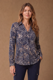 A woman modeling an EMRECO long-sleeve button-up blouse with a navy blue and beige abstract reptile or snakeskin-style pattern, paired with dark navy pants, against a solid warm brown studio background. Available to buy from mooreheads.co.uk 