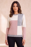 A woman wearing an Emreco colour-block knit sweater with cream, dusty pink, and light gray panels, paired with black pants, standing against a warm beige textured background.