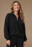 A woman wearing an EMRECO black long-sleeve quarter-zip sweatshirt and matching black sweatpants, standing against a plain beige studio background. Available to buy at Mooreheads Bessbrook.