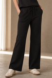 A person wearing EMRECO black wide-leg drawstring pants with side pockets, paired with a black top and white sneakers. Availableto buy from mooreheads.co.uk 