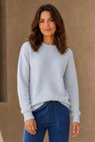 A woman wearing an Emreco light blue bubble textured crewneck sweater and blue jeans, standing against a neutral beige wall.