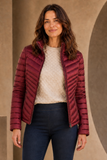 A woman wearing an Emreco quilted puffer jacket in burgundy, styled over a cream textured knit sweater and dark blue jeans, standing against a warm beige architectural background.