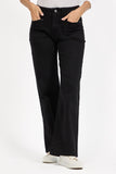 A person wearing MUDFLOWER black wide-leg jeans with hands in the front pockets, paired with a white top and white sneakers, photographed against a clean white studio background. Available to buy from W. Moorehead, Bessbrook.