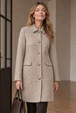 A woman wearing an Emreco mid-length brown herringbone coat with large buttons and flap pockets, standing indoors and holding a dark brown tote bag.