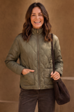 A smiling woman wearing an Emreco olive green quilted zip-front jacket with front pockets, paired with dark brown pants and carrying a dark brown shoulder bag, set against a warm brown textured background.